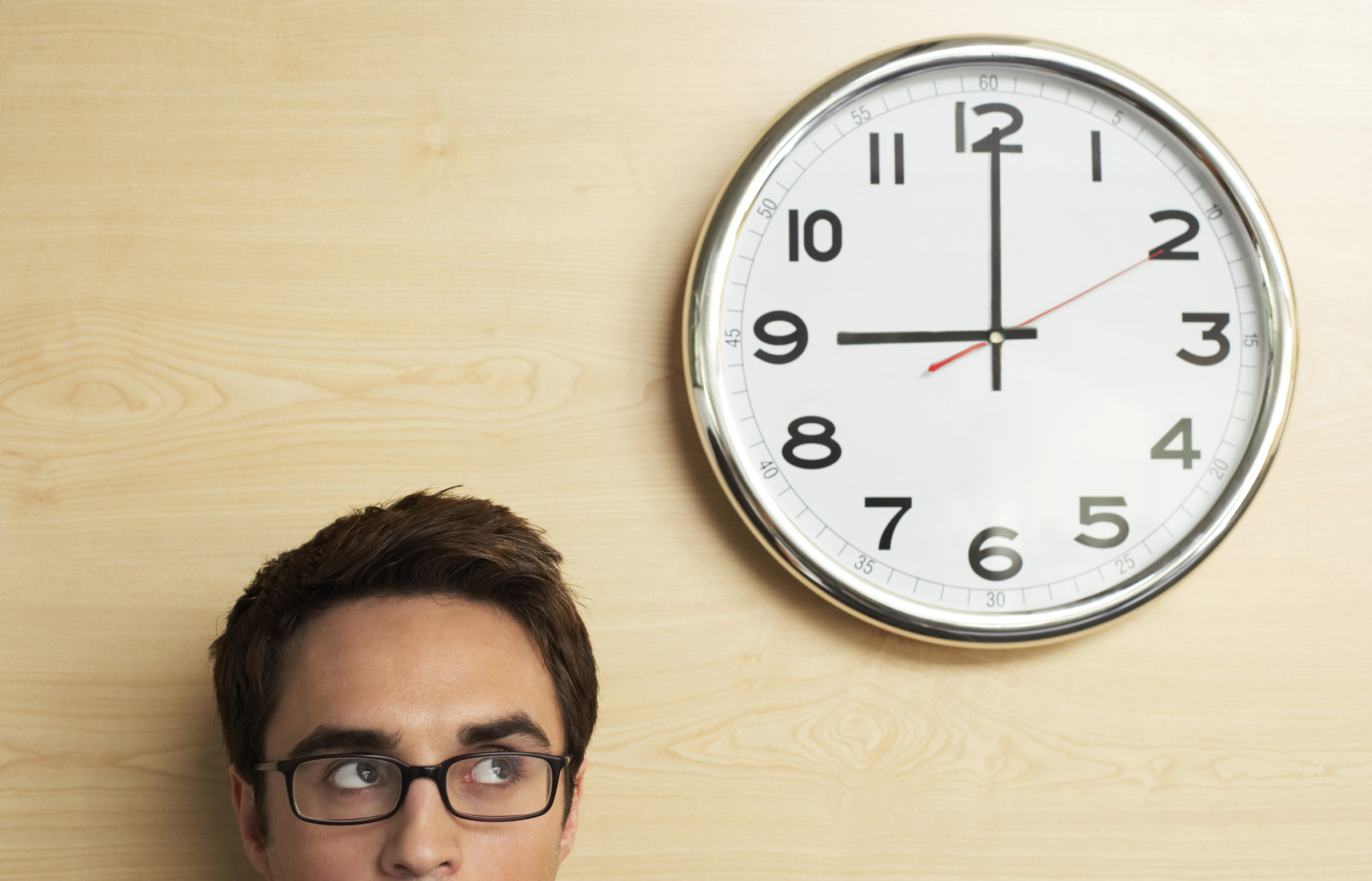 Headshot of young businessman wearing spectacles looking at clock on wooden wall in office